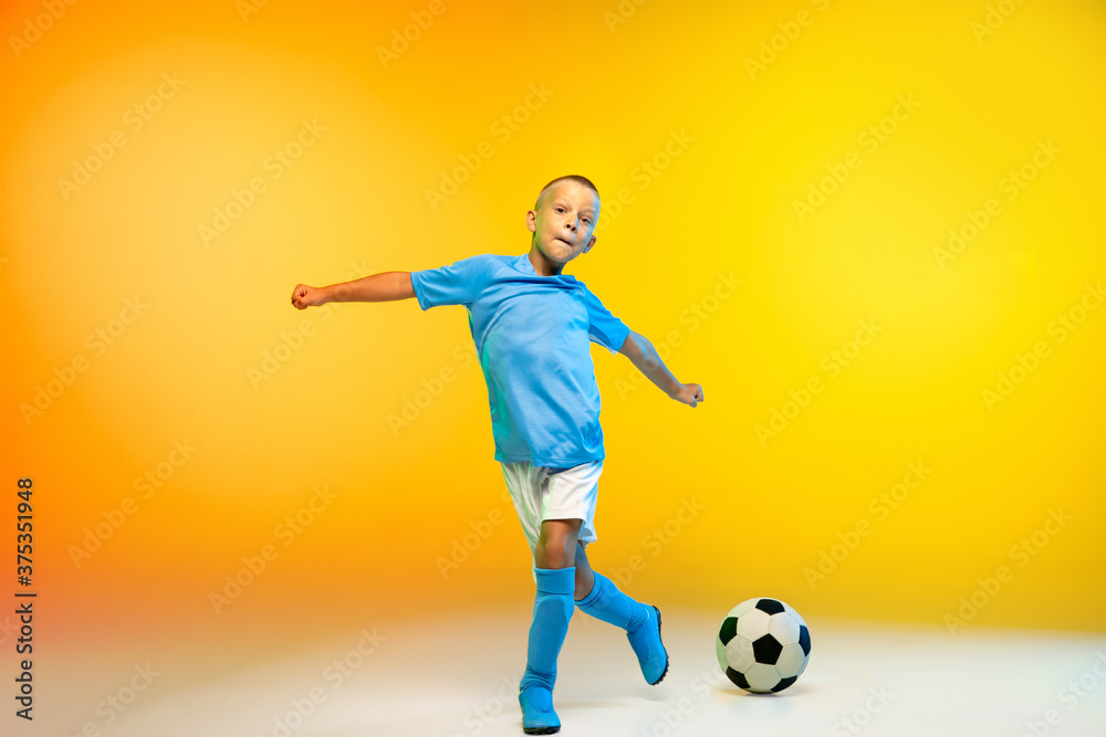 Attack. Young boy as a soccer or football player in sportwear practicing on gradient yellow studio background in neon light. Fit playing boy in action, movement, motion at game. Copyspace.