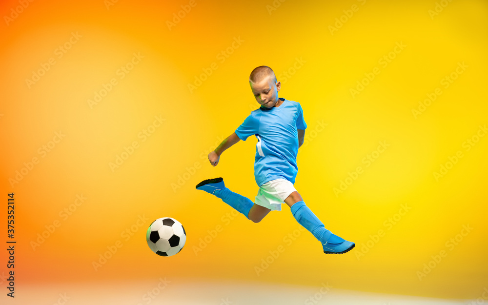 In flight. Young boy as a soccer or football player in sportwear practicing on gradient yellow studio background in neon light. Fit playing boy in action, movement, motion at game. Copyspace.