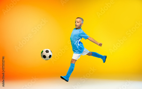 In flight. Young boy as a soccer or football player in sportwear practicing on gradient yellow studio background in neon light. Fit playing boy in action, movement, motion at game. Copyspace. © master1305
