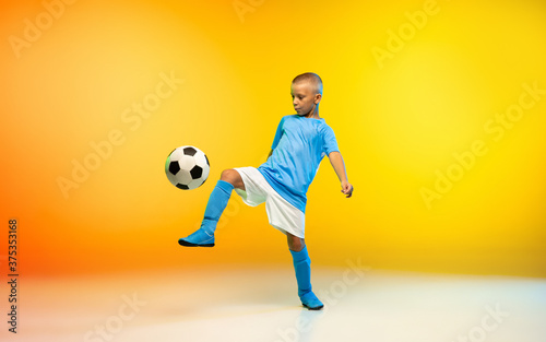 Fototapeta Naklejka Na Ścianę i Meble -  Kicking. Young boy as a soccer or football player in sportwear practicing on gradient yellow studio background in neon light. Fit playing boy in action, movement, motion at game. Copyspace.