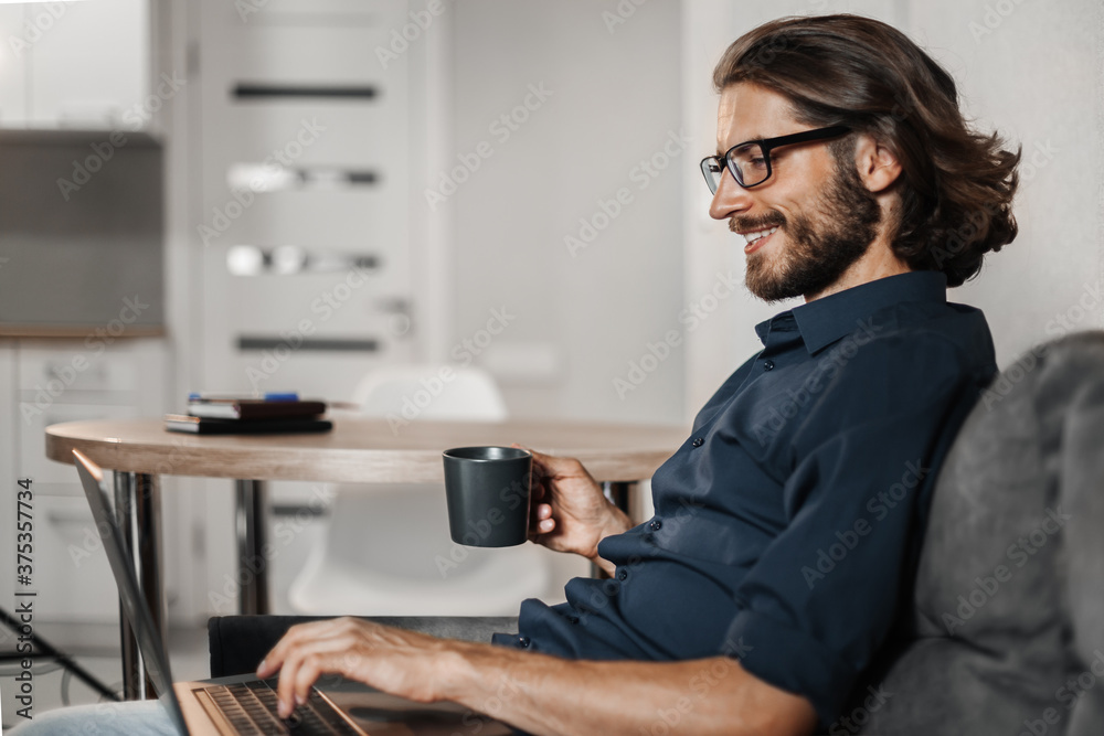 Mulody man with long hair works at home using laptop while sitting on sofa Mulody man with long hair works at home using laptop while sitting on sofa