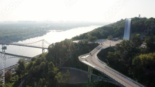 Kyiv Ukraine. view from Volodymyr hill to Dnipro river Parkovy Pedestrian, People's Friendship Arch and new Bicycle Bridge. Car traffic. Morning routine. Aerial vertical movement from right to left  photo