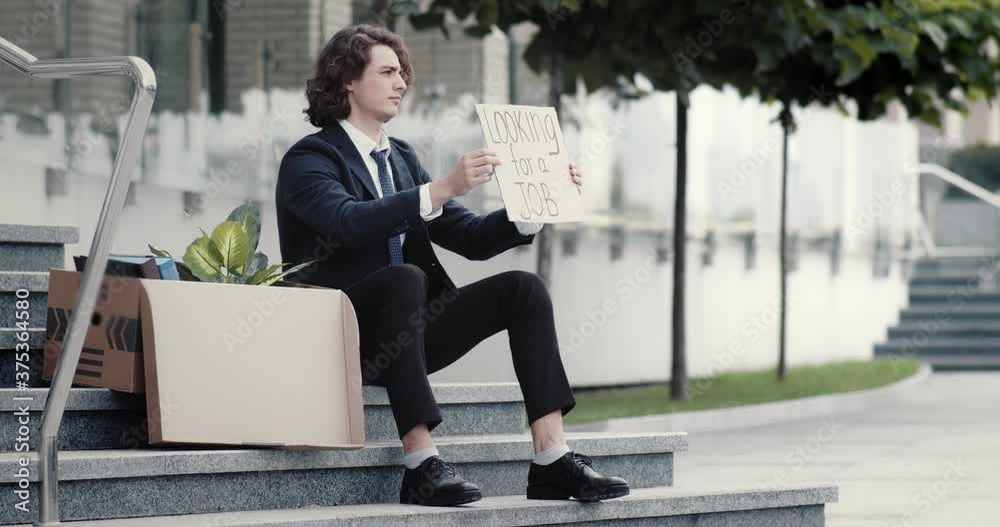 Unemployed businessman in despair sits on stairs outdoors next to a box with personal belongings, holding cardboard sign banner that says looking for a job. Fired office worker seeks help. Unemployed businessman in despair sits on stairs outdoors next to a box with personal belongings, holding cardboard sign banner that says looking for a job. Fired office worker seeks help.