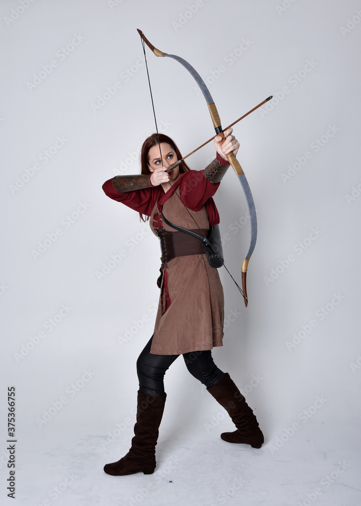 Full length portrait of girl with red hair wearing  brown medieval archer costume.. Standing pose holding a bow and arrow,  isolated against a grey studio background.