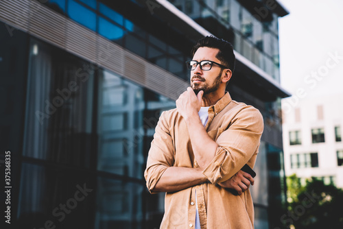 Pensive young male in casual wear and eyeglasses puzzled on smth standing on street,confident thoughtful businessman in spectacles looking away concentrated on ideas standing on urban settings