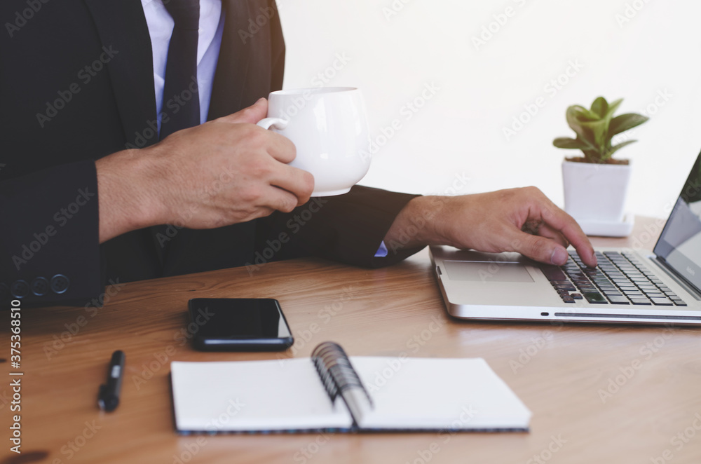 The businessman is working with a laptop, holding a mobile phone in the office, and drinking coffee. The businessman is working with a laptop, holding a mobile phone in the office, and drinking coffee.