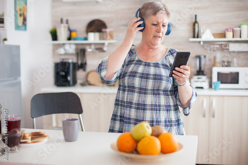 Relaxed senior woman listening music on headphones during breakfast in kitchen. Elderly person dancing, fun lifestyle with modern technology