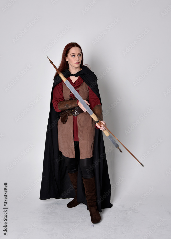 Full length portrait of girl with red hair wearing medieval archer costume with black cloak. Standing pose with back to the camera holding a bow and arrow, isolated against a grey studio background. Full length portrait of girl with red hair wearing medieval archer costume with black cloak. Standing pose with back to the camera holding a bow and arrow, isolated against a grey studio background.