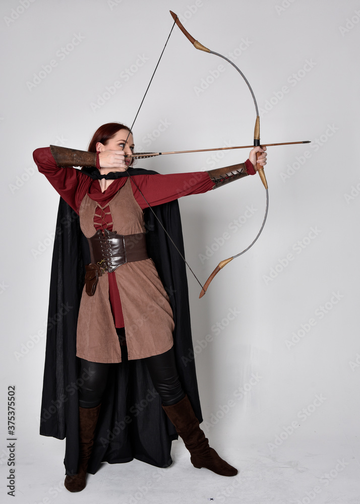 Full length portrait of girl with red hair wearing medieval archer costume with black cloak. Standing pose with back to the camera holding a bow and arrow,  isolated against a grey studio background.