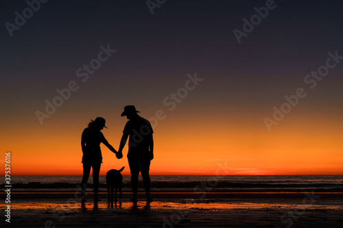 Silhouette of unrecognisable young couple holding hands and a dog in front of a glowing sunset on Australian beach.