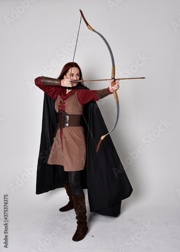 Stampa su tela Full length portrait of girl with red hair wearing medieval archer costume with black cloak