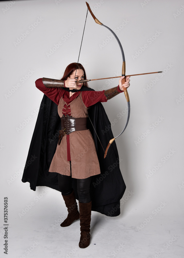 Full length portrait of girl with red hair wearing medieval archer costume  with black cloak. Standing