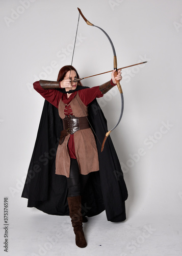 Full length portrait of girl with red hair wearing medieval archer costume with black cloak. Standing pose with back to the camera holding a bow and arrow, isolated against a grey studio background.