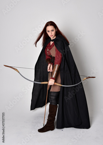 Fototapet Full length portrait of girl with red hair wearing medieval archer costume with black cloak