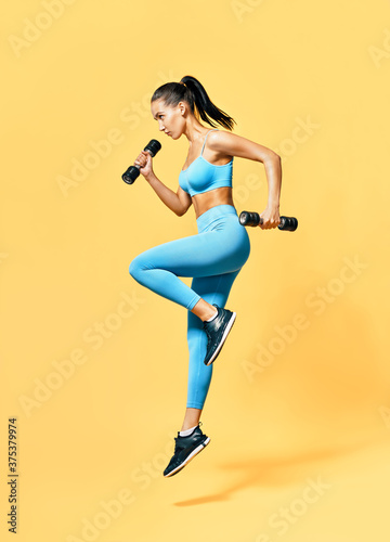 Fototapeta Naklejka Na Ścianę i Meble -  Sporty woman in sportswear jumping with dumbbells on yellow background