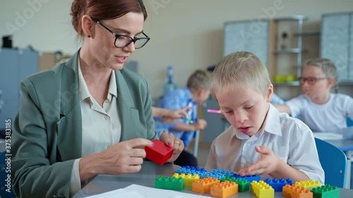 specialized school, down syndrome boy plays and counts use colorful constructor sitting with teacher at the desk during lesson