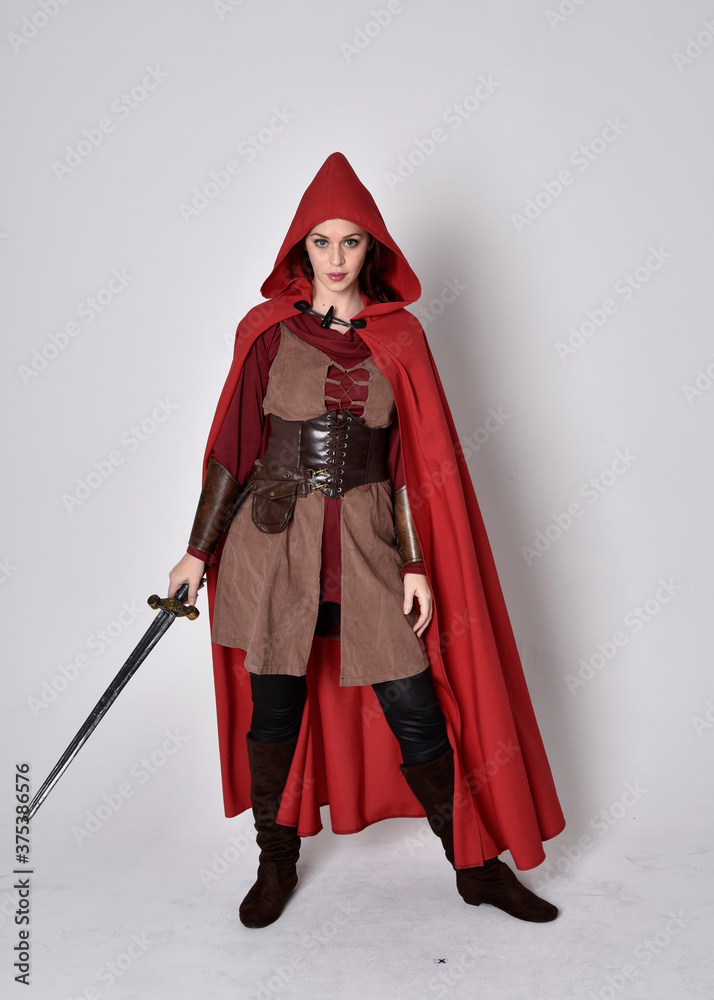 Full length portrait of girl wearing medieval costume and red cloak. Standing pose holding a sword,  isolated against a grey studio background.