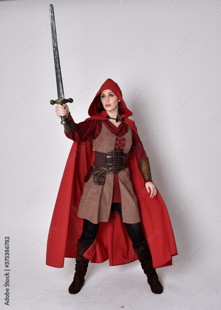 Full length portrait of girl wearing medieval costume and red cloak. Standing pose holding a sword, isolated against a grey studio background. Full length portrait of girl wearing medieval costume and red cloak. Standing pose holding a sword, isolated against a grey studio background.