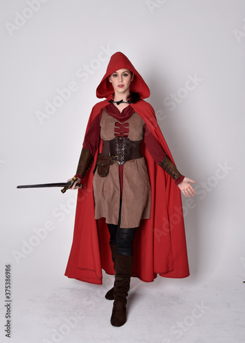 Full length portrait of girl wearing medieval costume and red cloak. Standing pose holding a sword, isolated against a grey studio background.