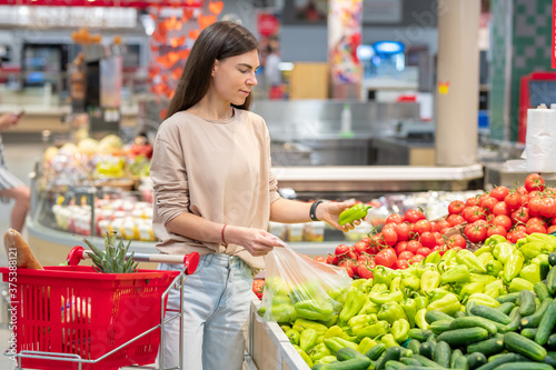 Portrait of young adult woman standing in supermarket with shopping cart choosing sweet peppers © pressmaster