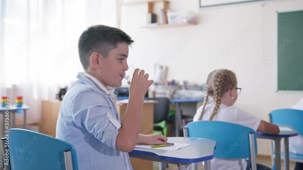 schoolboy drinking water while sitting at a desk in the classroom during a lesson on background of classmates, elementary school schoolboy drinking water while sitting at a desk in the classroom during a lesson on background of classmates, elementary school