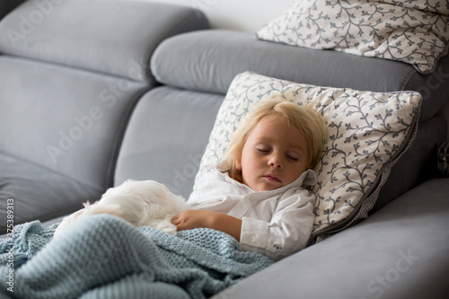 Sick child, toddler boy lying on the couch with his white puppy in living room with fever, resting