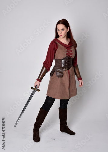 Full length portrait of girl wearing medieval costume. Standing pose holding a sword, isolated against a grey studio background.