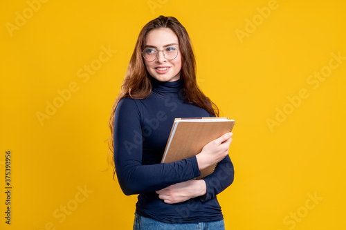 Teacher portrait. Higher education. Cheerful smart woman in glasses standing with books smiling isolated on yellow copy space background. Academic degree. Professional training course.