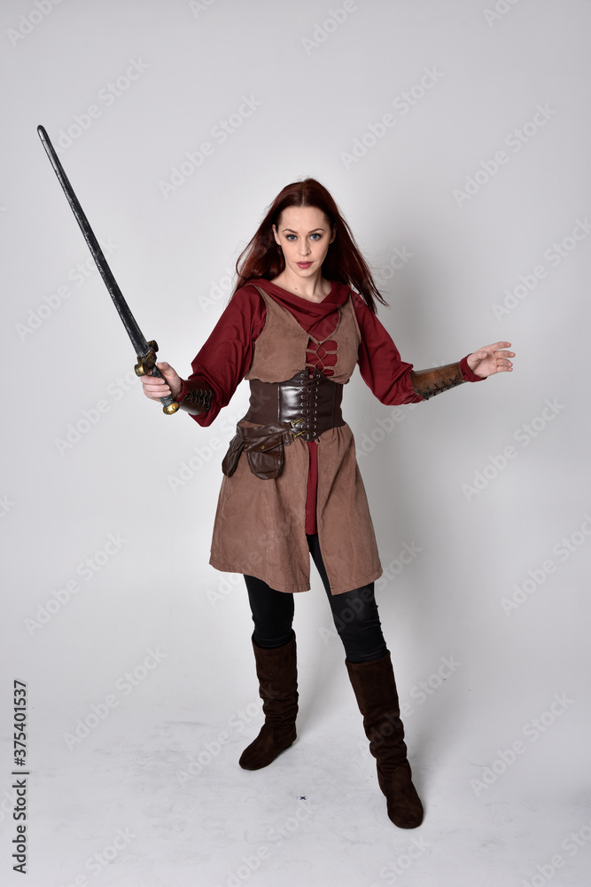 Full length portrait of girl wearing medieval costume. Standing pose holding a sword, isolated against a grey studio background. Full length portrait of girl wearing medieval costume. Standing pose holding a sword, isolated against a grey studio background.