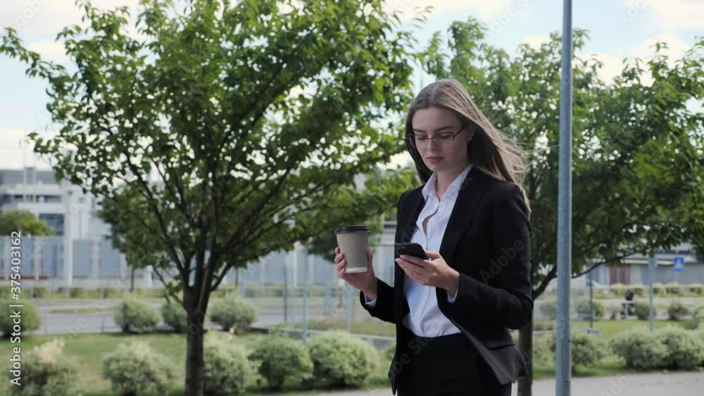 Beautiful businesswoman goes to a meeting while typing a message and holding a coffee on a background of green trees. Beautiful businesswoman goes to a meeting while typing a message and holding a coffee on a background of green trees.