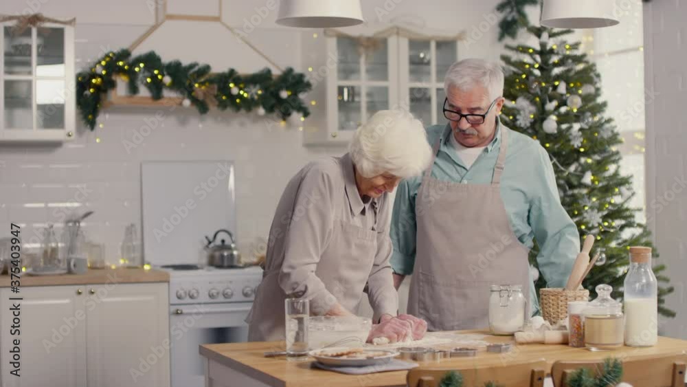 Medium shot of elderly woman and man in aprons baking together in kitchen on Christmas day Medium shot of elderly woman and man in aprons baking together in kitchen on Christmas day