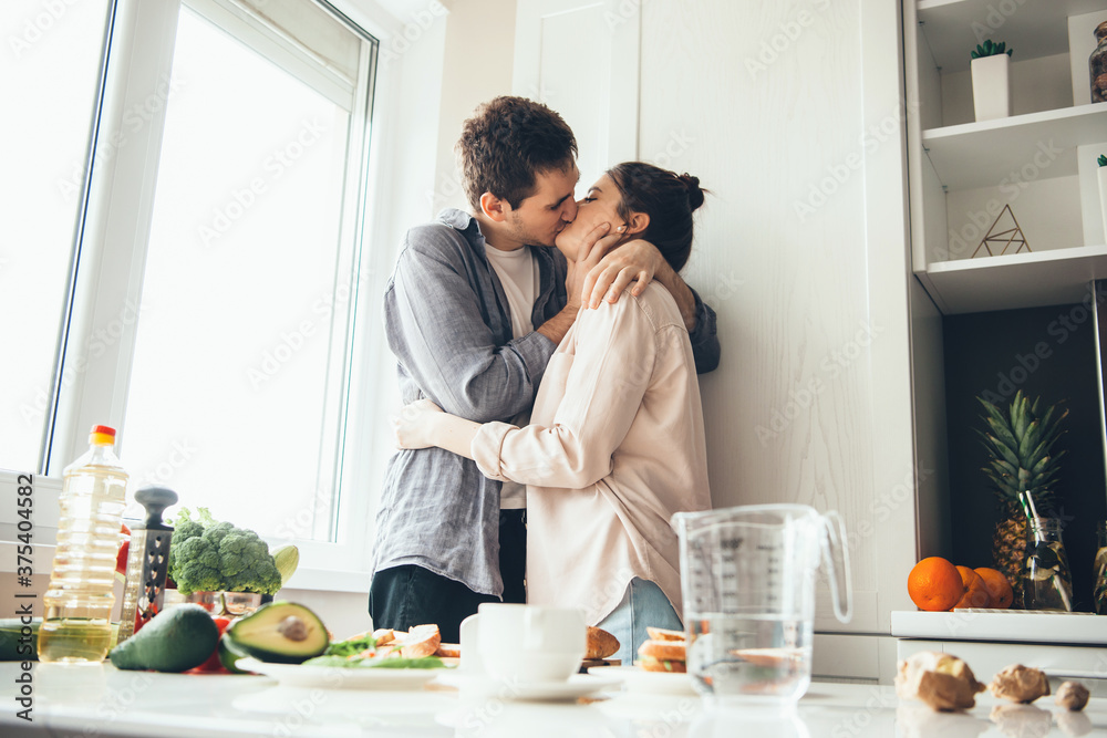 Caucasian couple kissing in the kitchen while preparing food near the window Caucasian couple kissing in the kitchen while preparing food near the window