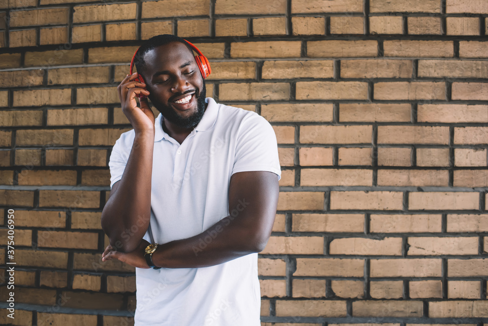 Cheerful dark skinned hipster guy in red headphones satisfied with sound of favorite music songs, African American male millennial with closed eyes enjoying audio podcast at promotional background