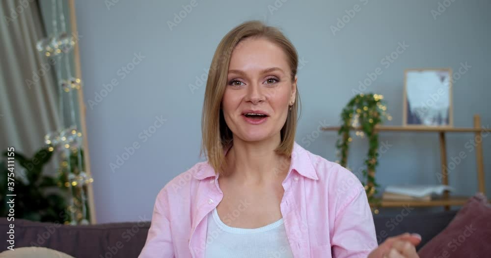 Beautiful girl speaking and smiling while looking to camera and sitting on sofa. Beauty female vloger moving hands while recording new video episode for her vlog. Concept of vlogging. Beautiful girl speaking and smiling while looking to camera and sitting on sofa. Beauty female vloger moving hands while recording new video episode for her vlog. Concept of vlogging.
