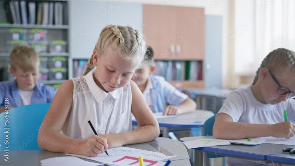 portrait of happy pupil girl doing classwork sitting at a desk on background of classmates at elementary school portrait of happy pupil girl doing classwork sitting at a desk on background of classmates at elementary school