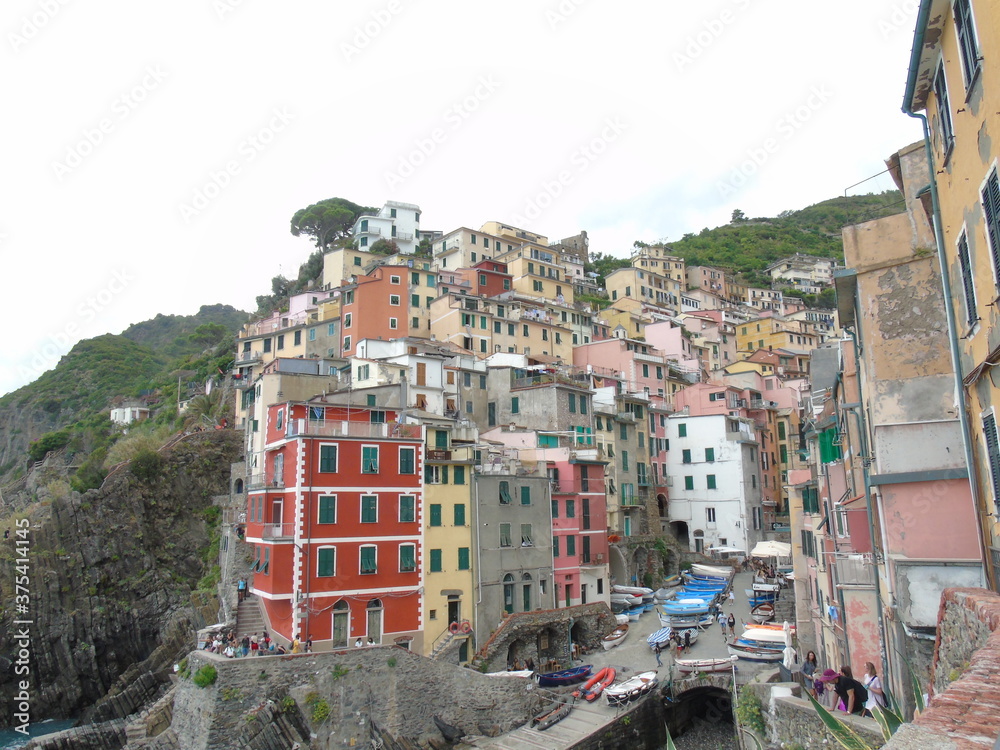 Fototapeta premium Riomaggiore, Italy - 08/31/2020: Beautiful photography of the countryside from Cinque Terre, Italy. Grey sky, colourful houses, grape coltivation from the mountains, and some small waves from the sea.