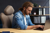 Young handsome curly smiling man with long hair working in home office with laptop. Business portrait of handsome manager sitting at workplace. Studying online, online courses. Business concept.