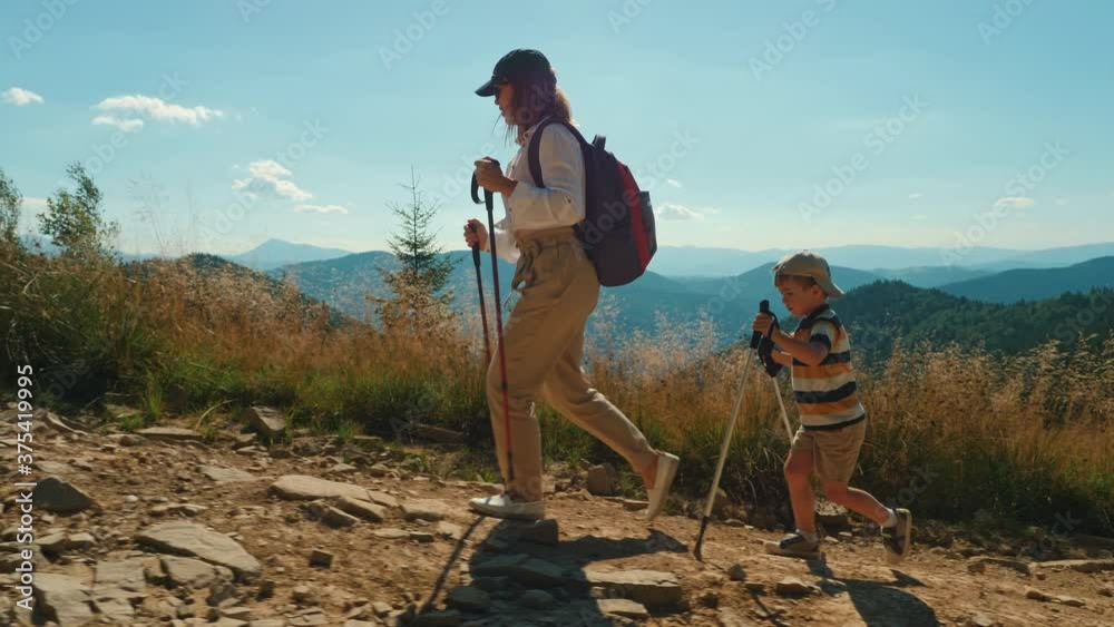 A young active mother with her little son climbing mountain. Woman and child with trekking poles and backpack hike together along a rocky path