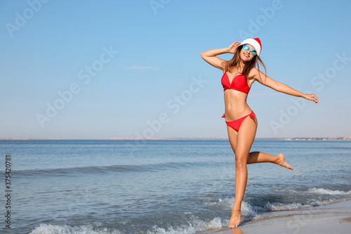 Young woman wearing Santa hat and bikini on beach, space for text. Christmas vacation