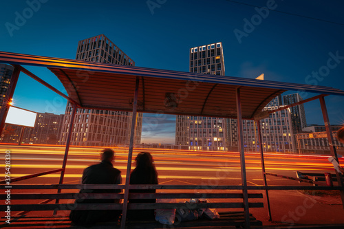 An illuminated city street at the night. The bus stop with silhouettes of a couple, sitting on the bench, in front of bright light trails, from moving cars.
