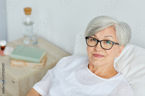 Portrait of Caucasian mature woman wearing eyeglasses recovering in hospital ward looking at camera