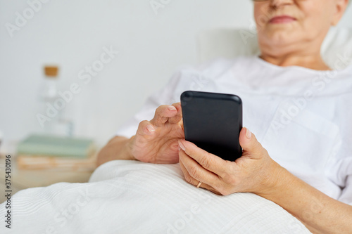 Modern aged woman wearing white clothes relaxing on bed in hospital ward texting messages using her smartphone