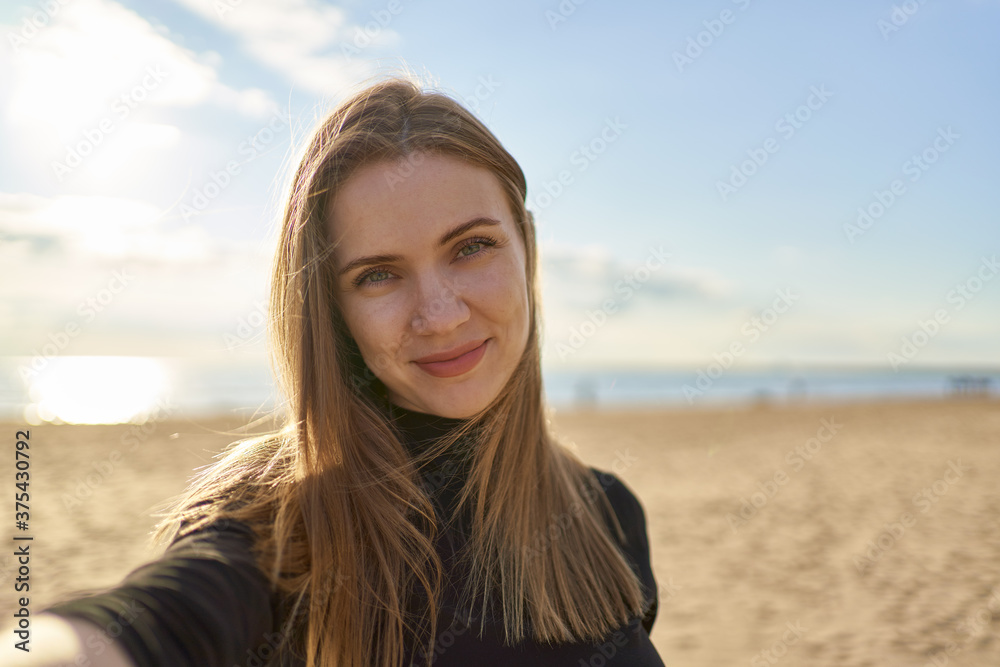 Pretty female with long hair looking at camera and smiling in sunny evening in ocean or sea coastline. Woman takes selfie photo on mobile phone on sandy beach in summer or autumn