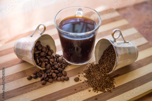 On a wooden plank in the middle is a cup of brewed coffee, the other two cups are filled with whole, roasted beans and instant coffee, and they are scattered on the plank.