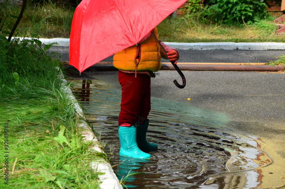 little funny cute girl walks in the rain with a red umbrella, in green rubber boots through the puddles. Laughs, has fun. girl wearing yellow waterproof coat playing on a warm autumn or summer day