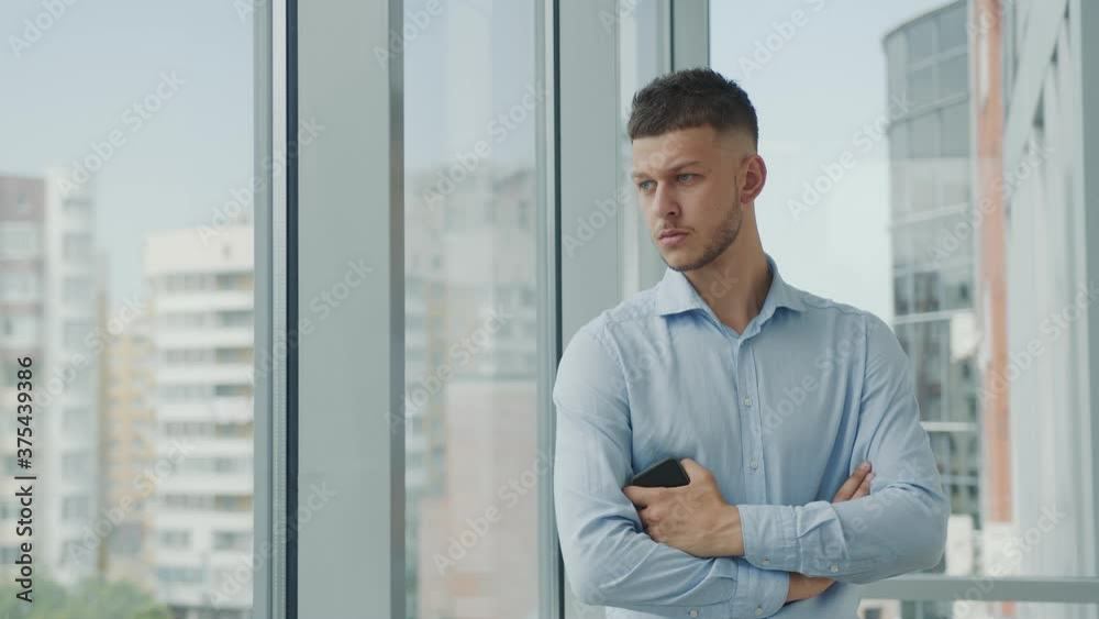Portrait of a young businessman in the office standing by the window with a pensive look thinking business strategy, and looking at messages in a mobile phone. Portrait of a young businessman in the office standing by the window with a pensive look thinking business strategy, and looking at messages in a mobile phone.