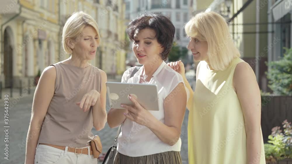 Portrait of three cheerful mid-adult women standing in sunlight on city street and using tablet. Happy Caucasian tourists choosing place for sightseeing in foreign town. Summer tourism concept. Portrait of three cheerful mid-adult women standing in sunlight on city street and using tablet. Happy Caucasian tourists choosing place for sightseeing in foreign town. Summer tourism concept.