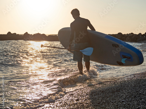 Photography Man with SUP and paddle on beach go surfing at golden sunset background