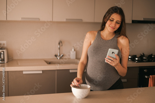 Pregnant beauty young woman in kitchen watching the phone (ID: 375448956)