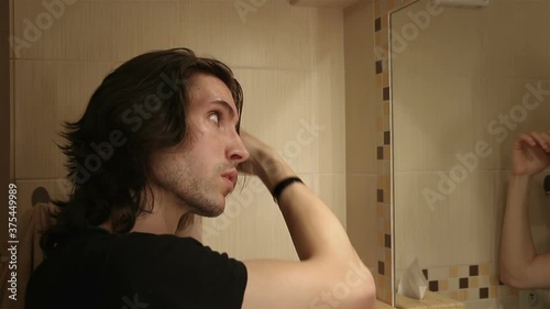 Portrait of young man brushing long layered dark brown hair in bathroom standing in front of mirror, holding comb, face profile photo